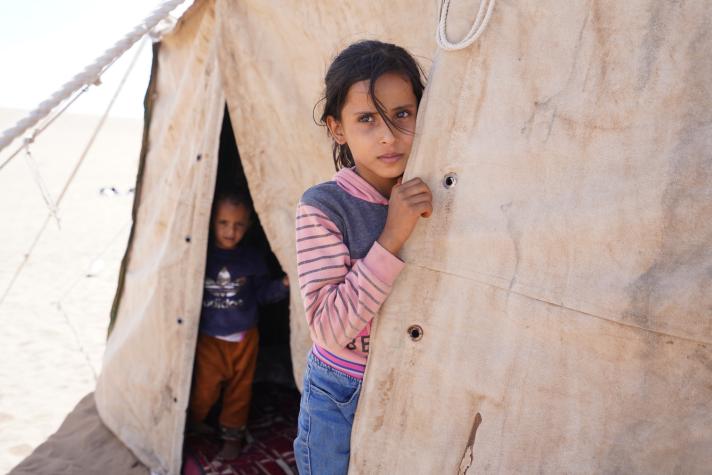 A girl standing at the entrance of a shelter tent. In the background a little boy standing in another opening of the shelter tent.