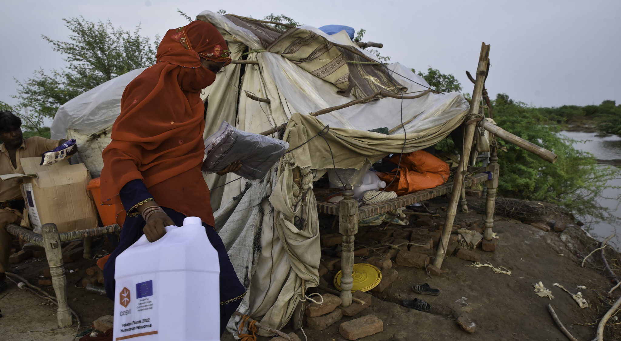 Woman carrying a water container, in the background a make-shift shelter and a river.