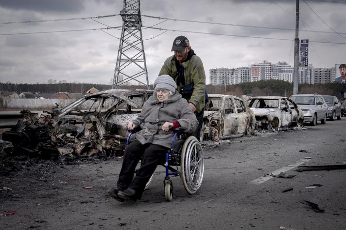 Eldery in a wheel chair being helped along a street with burnded out cars along the side