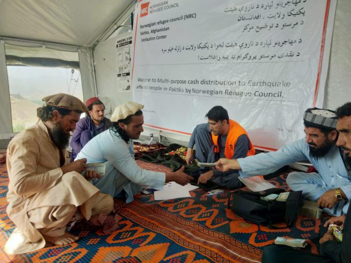 Group of men sitting on a carpet in a tent receiving their cash assistance