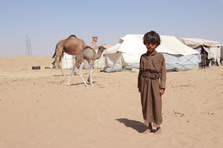 Child standing in the desert, on the background their tent and a camel.