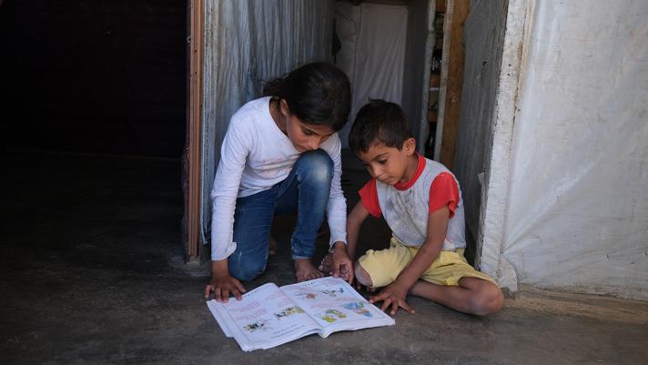 2 children sitting on the ground reading a schoolbook together.