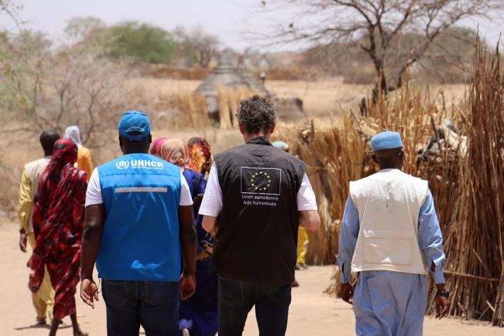 Aid workers and local people seen from the back, walking on a sandy road.