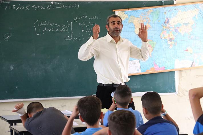 Hani Bdair, a geography teacher, in front of a class. Behind his back is a blackboard and a map of the world.