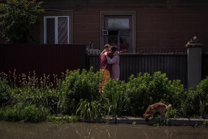 2 persons hugging eachother at the side of a flooded street. A dog drinking water from the street.