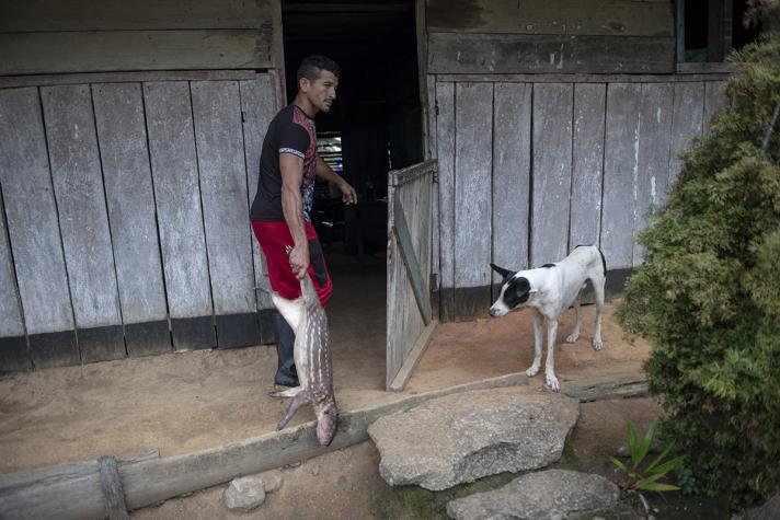 A man, holding a dead animal, entering his house. At the left a dog watching.