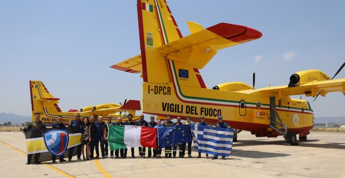 Teams of firefighters holding different EU flags, standing in front of 2 water-bombing planes.