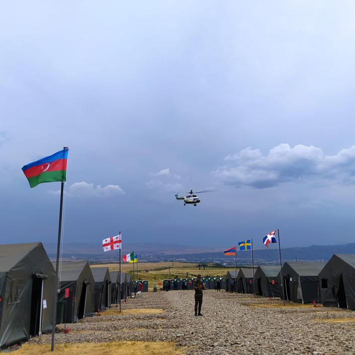View of a field, on both sides tents have been put up each carrying a national flag. A helicopter is in the air.