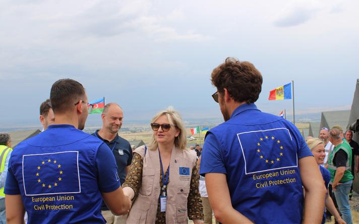 A woman shaking hands with 2 people, seen from the back, wearing European Union Civil Protection jackets.