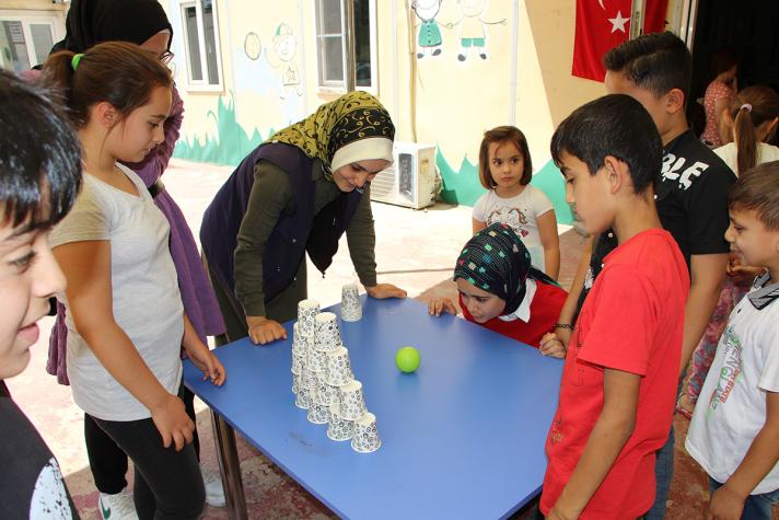 Children playing a game on a table. A ball is thrown towards a pile of cups placed on top of each other. Around other children and a teacher.