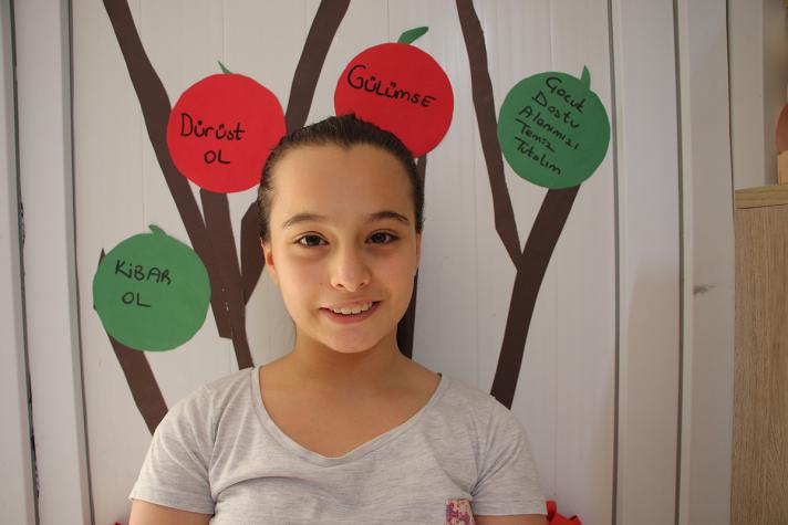 Photo of Ece in front of a colourful collage representing a tree with red and green circles with text written on it.