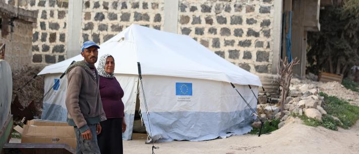 A woman and a man standing next to a shelter tent. In the background a brick wall.