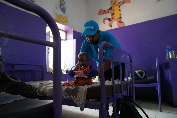 Health and Nutrition Officer Mohamed Almugtba Khider feeds 16 months old Mahdi while sat on a bed.