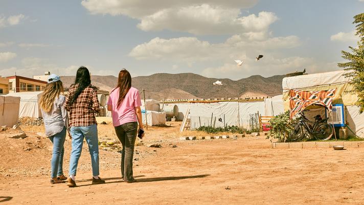 3 youngsters walking towards a settlement in a sandy area. In the background some mountains.