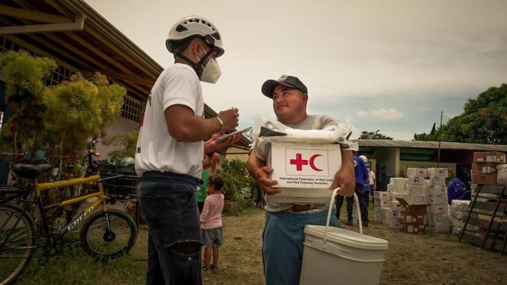 A person receiving a box with emergency goods from an aid worker wearing a helmet and a mask.