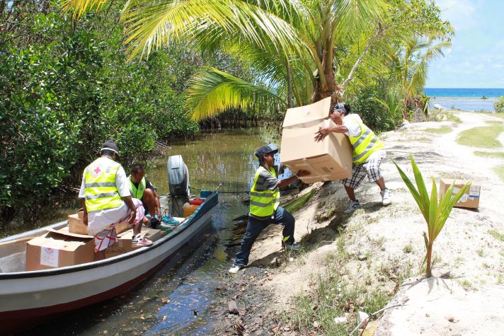 People at the right side unloading big boxes from a longboat laying in a river on the left side.