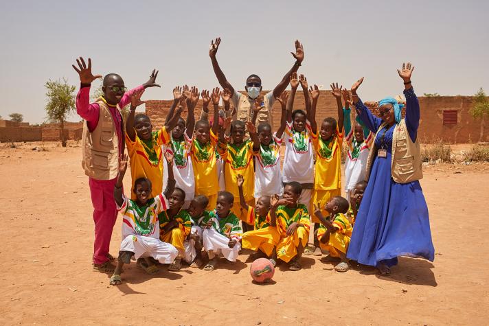 A group photo of children in similar clothing flanked by some adults. The all hold their hands high like celebrating.