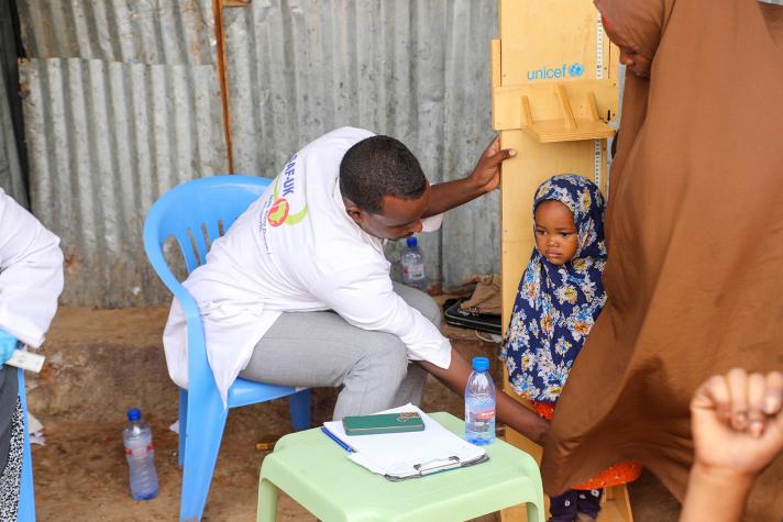 A child’s height is being measured by a health worker who is seated on a chair.