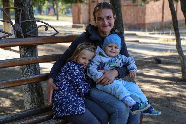 Iryna sitting on a bench in a park while holding her 2 children close to her.