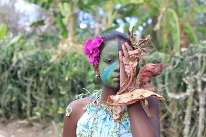A girl with a flower in her hair, her face coloured in Green and holding her hand against the right side of her face to make it invisible.