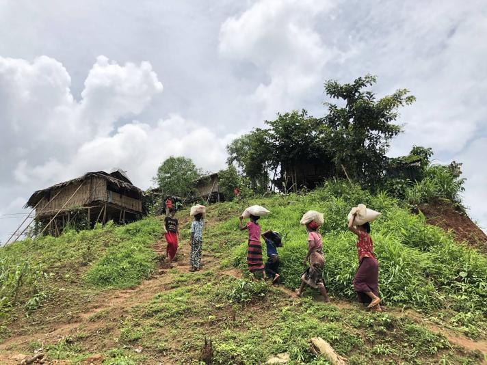 5 women walking uphill towards a house while carrying goods on their heads