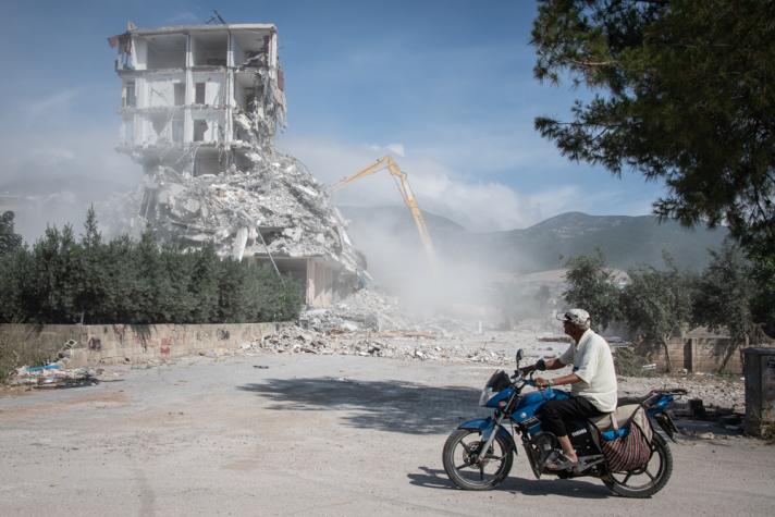 A man on a motorbike in front. In the back an appartment building being demolished.