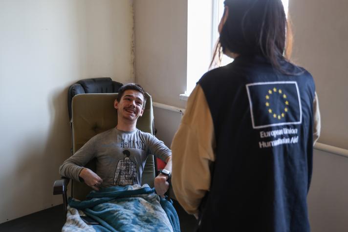 Ihor seated while talking to an aid worker, seen from the back wearing a jacket with a print of the EU flag on it.