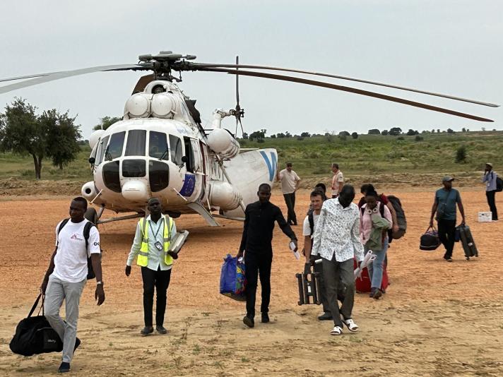 A crowd of people with luggage on the air strip, in the back a helicopter.
