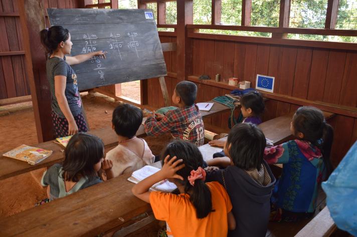 View of a classroom in a wooden building. Some pupils and a teacher in front of them pointing to something at the blackboard.