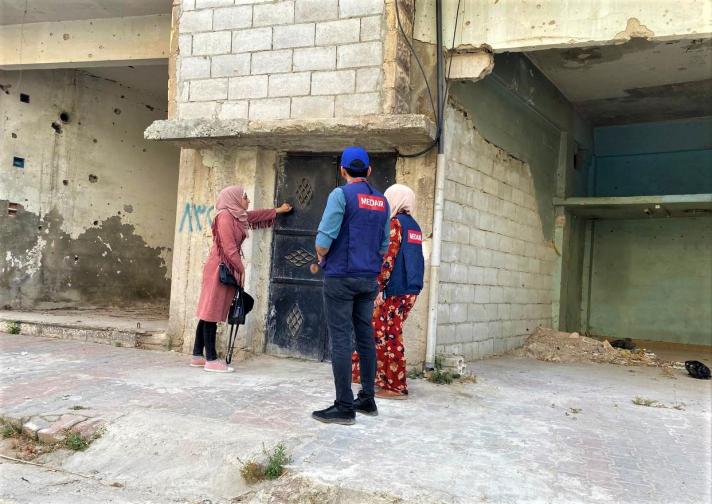 Medair's health team stands at the doorstep of what’s left of Rabiea's home. Rabiea is pointing towards some damage.