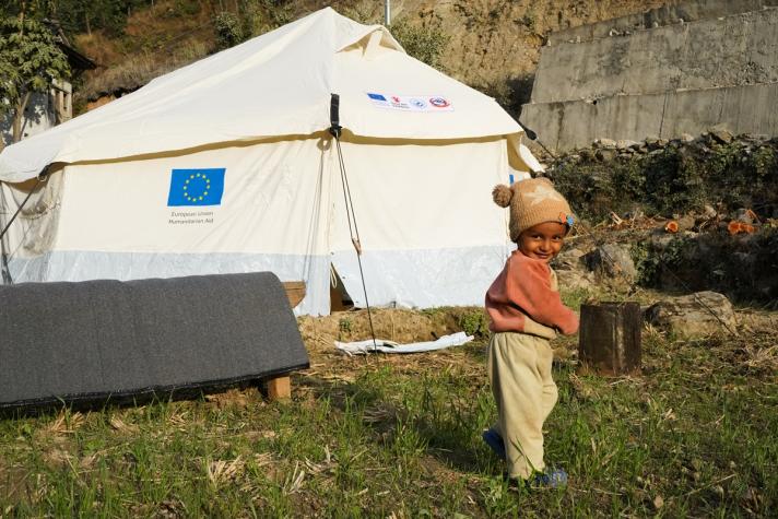 A child looking into the camera, in the background a shelter tent.