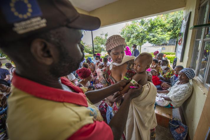 A woman holding her young child in her arms. A health worker measuring the child's arm.