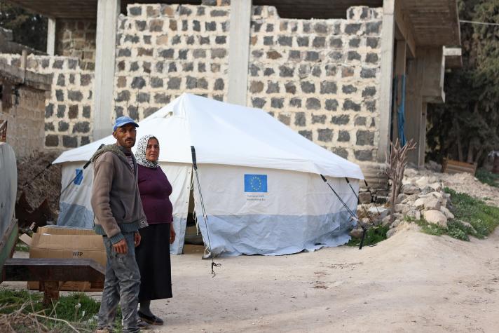 An elderly couple in front of a shelter tent which stands in front of their damaged building.