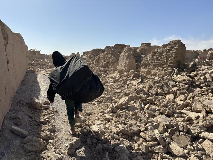 A person carrying a bag on the shoulder while walking inbetween destroyed buildings.