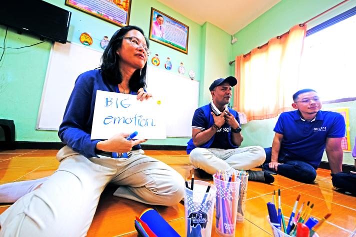 An aid worker holding up a paper with the text 'big emotions'. Pencils are on the floor and 2 other people in the background.