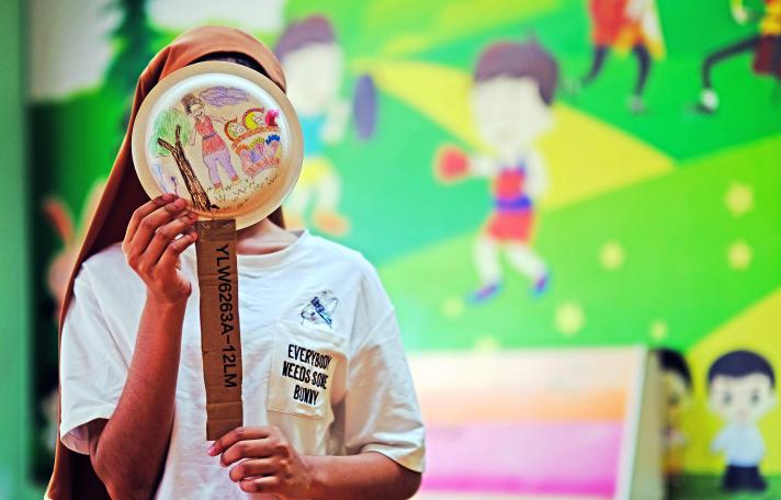 A girl holding a paper plate, with drawings on it, in front of her face. In the background a colourful wall with drawings of children playing.