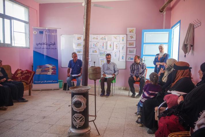 Group of people sitting alongside the walls of a room. In the middle a coal heater.