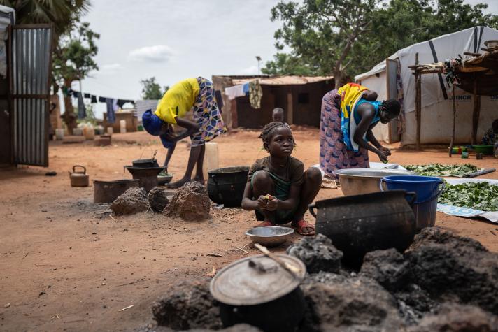 View of a street in a village. In the front a woman preparing a meal in a big bowl.