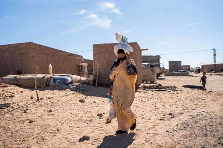 A woman carrying goods on her head, in the background some buildings and a child.