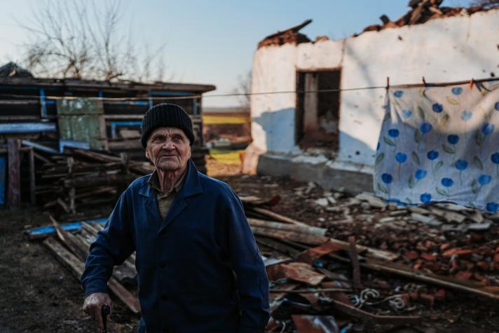 An elderly man standing in his garden, a destroyed house in the background