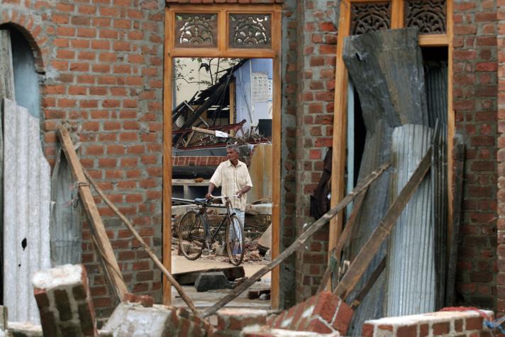 A person holding a bicycle standing in front of a house destroyed