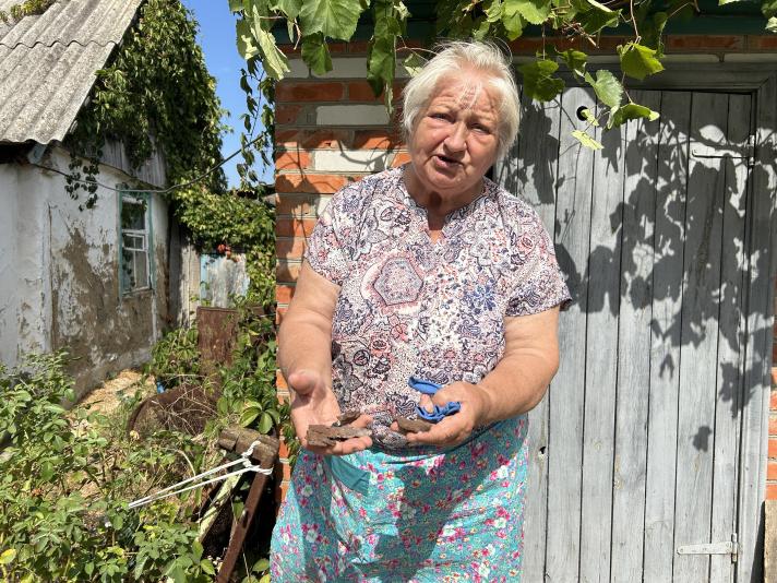 Paraskoviia, standing in her garden, showing some bomb parts she holds in her hands.