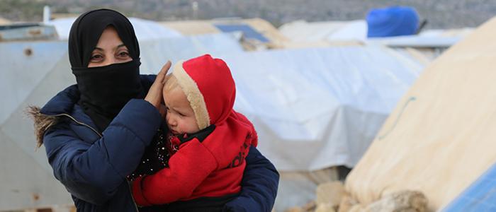 Woman holding child in her arms with on the background a refugee camp