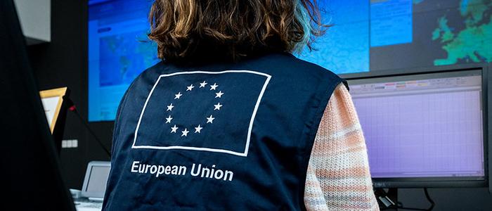 View of the back of a person in front of a screen wearing a jacket with the EU flag and text Humanitarian Aid