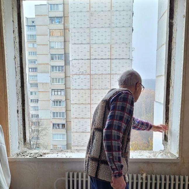 Photo of Yevhen in front of a broken window. In the background an apartment building.