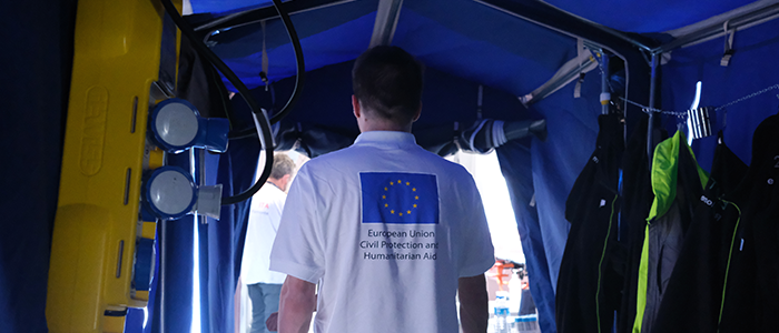 View from inside a field hospital to outside. In the doorway a person wearing a white t-shirt with the EU flag printed on it.