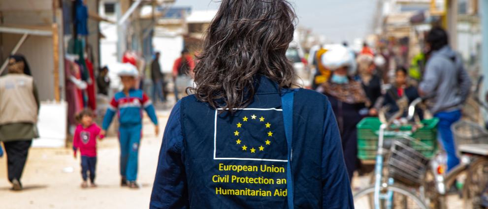 Aid worker in a street, seen from the back. A EU flag with text shown on the back of her jacket.
