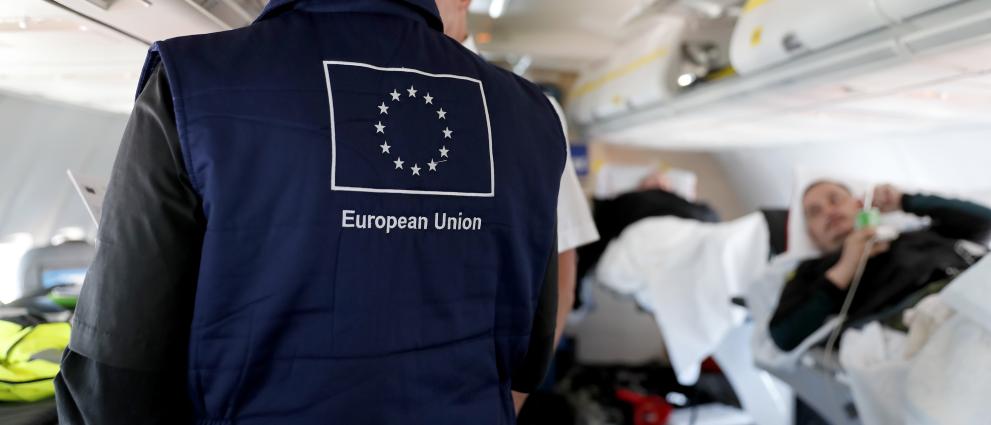 View inside a plane, a person wearing a vest with the EU flag. At the right a patient laying in a bed.