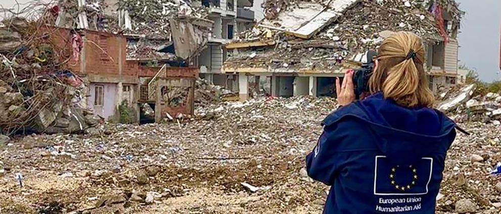An aid worker seen from the back taking pictures of a destroyed building.