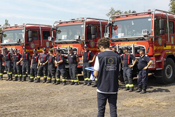 Several firefighters line up in front of two fire trucks, accompanied by EU staff.